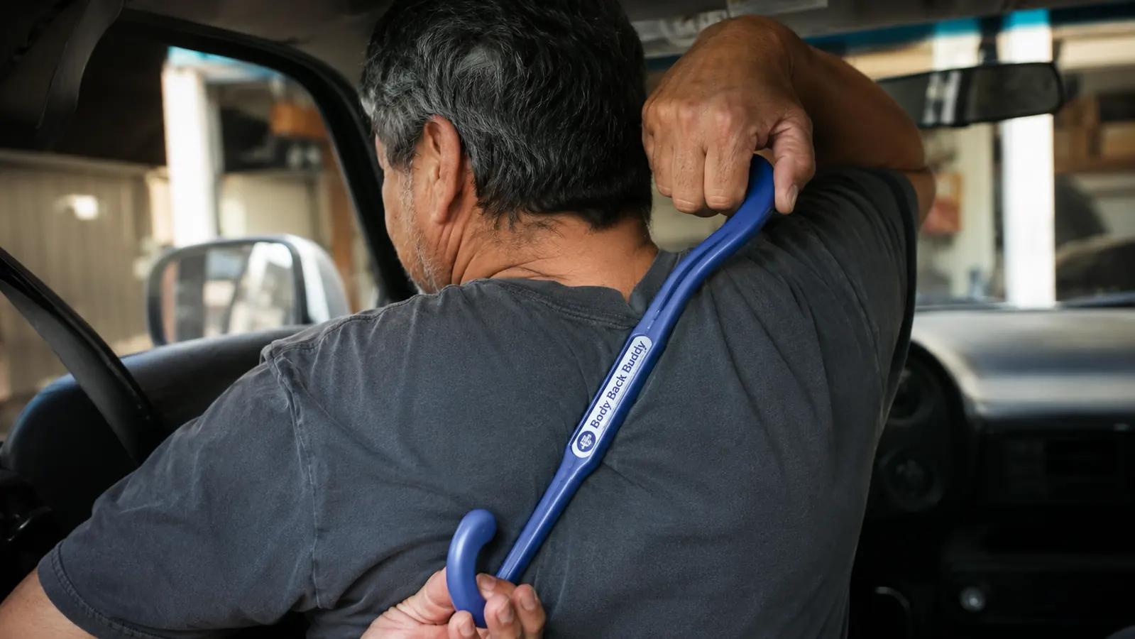 Driver's hand using a two-hook massage cane to press into the muscle between his shoulder blade and spine