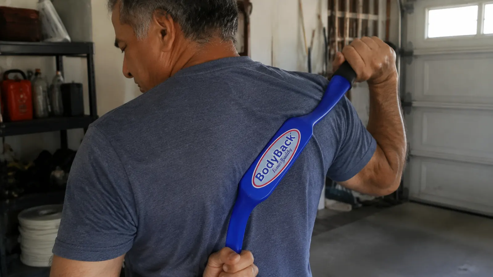 Close-up of a hand awkwardly gripping a Body Back Buddy massage cane at the wrong angle against the upper back, showing the learning curve