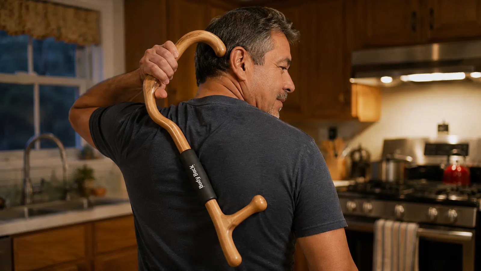 A man at his kitchen counter using a hooked wooden massage cane over his shoulder to reach a knot in his upper back