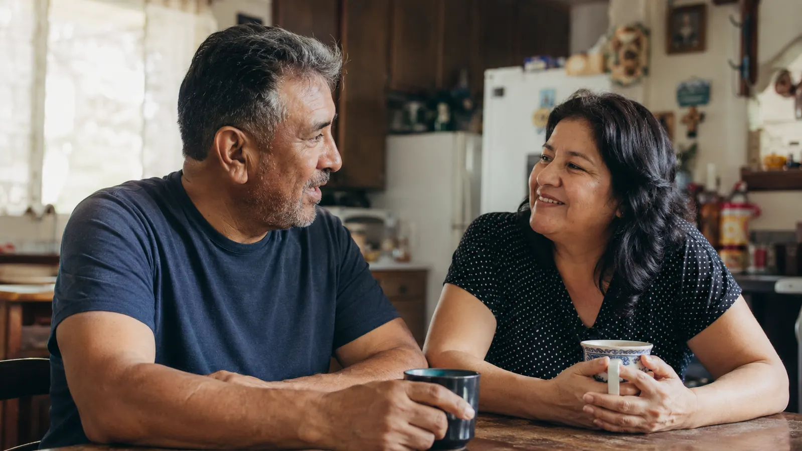 A man and woman sitting together at a small kitchen table with coffee mugs, warm morning light through a window